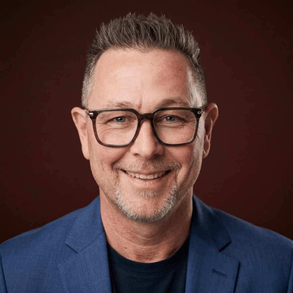 marc-ulrich-aivalley Mann mit Brille und kurzen Haaren, lächelnd, trägt einen blauen Blazer vor einem dunklen Hintergrund.